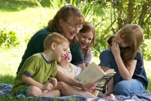 A woman and her three kids sitting on a blanket, reading outside.
