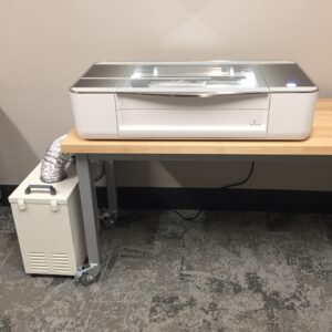 White Glowforge machine sitting on a table. Attached is a filter.