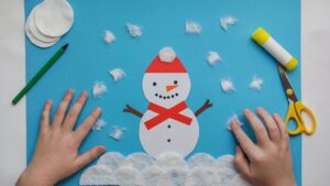 A pair of hands making a snowman on blue construction paper. Multiple pieces of cotton balls can be seen as snow.